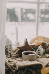 Warm cup of tea, candles, lights, little christmas house and tree, wooden star on cozy blanket on windowsill. Winter hygge. Atmospheric Christmas background. Cozy home on snowy day