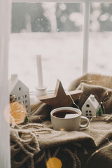 Winter hygge. Warm cup of tea, candles, lights, little christmas houses and trees, wooden star on cozy blanket on windowsill. Cozy home. Atmospheric snowy day. Christmas background