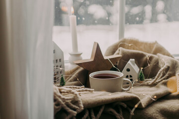 Fototapeta na wymiar Cozy home at snowy day. Warm cup of tea, candles, lights, little christmas houses and trees, wooden star on cozy blanket on windowsill. Winter hygge. Atmospheric scandinavian mood