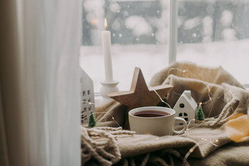 Winter hygge. Warm cup of tea, candles, lights, little christmas houses and trees, wooden star on cozy blanket on windowsill. Cozy home. Atmospheric snowy day. Christmas background