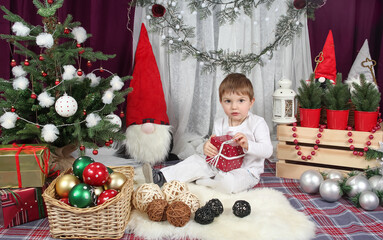 Portrait of a 3-year-old boy sitting under the Christmas tree with a New Year's gift in his hand, surrounded by fabulous Scandinavian gnomes in a red hat