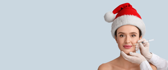 Fototapeta na wymiar Young woman in Santa hat receiving filler injection in lips against light background with space for text