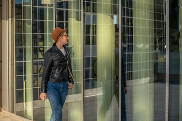 30s woman with short blonde hair in leather jacket and trendy hat looking on her reflection in mirror building in Barcelona city