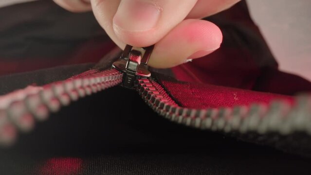 Hand of man fastens metallic zipper of dark-colored jacket under red artificial light of studio room pull out extreme closeup
