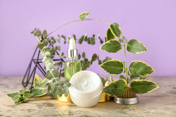 Jar of cream with cosmetic dropper bottle and ikebana on table near lilac wall