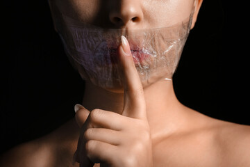 Young woman with taped mouth showing silence gesture on dark background, closeup. Censorship concept - obrazy, fototapety, plakaty