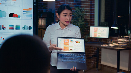 Asian employee presenting analytics research, showing statistics on laptop screen. Businesswoman presentation, diverse team discussing graphs, planning sales strategy in coworking space