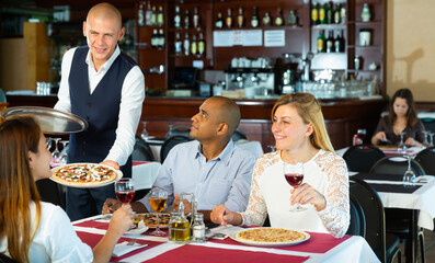Fototapeta na wymiar Portrait of smiling waiter serving delicious pizza to positive multiracial group of people in restaurant