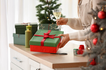 Woman with beautiful Christmas gift box and glass of champagne at table in kitchen