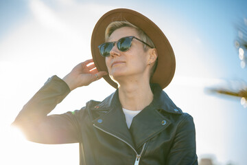 30s woman with short blonde hair in leather jacket and trendy hat sitting on a bench with palm trees on background in Barcelona city