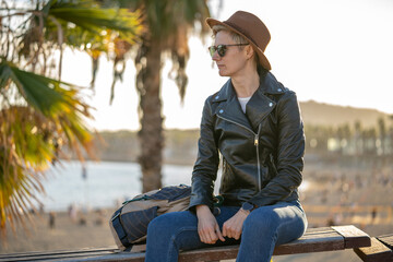30s woman with short blonde hair in leather jacket and trendy hat sitting on a bench with palm trees on background in Barcelona city