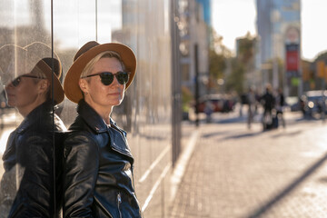 30s woman with short blonde hair in leather jacket and trendy hat standing next to the modern building in Barcelona