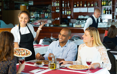 Positive waitress bringing ordered pizza to friends