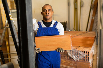 Focused Latino wearing blue coverall working in building materials hypermarket, arranging red bricks on display