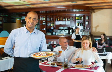 Obraz na płótnie Canvas Smiling waiter holding serving tray with pizza at restaurant with customers his behind