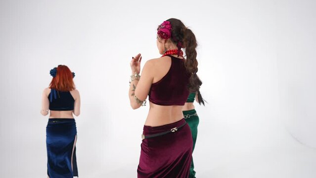 Three slim women spinning in slow motion shaking hips and chest at white background. Confident talented Caucasian belly dancers performing Middle Eastern dance