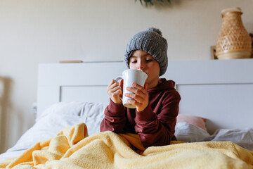 Child wearing grey warm hat sits on the bed under warm blanket and drinks hot tea