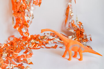 Toy dinosaur with long neck on white background with shiny orange and silver party streamers. Cute...