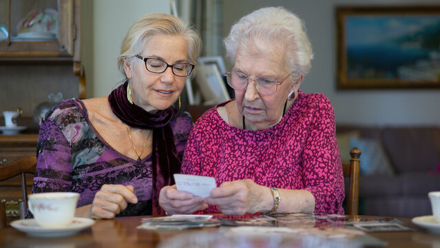 Senior elderly smiling happy woman looking at old photos having tea and remembering memories with daughter at the dining room table.