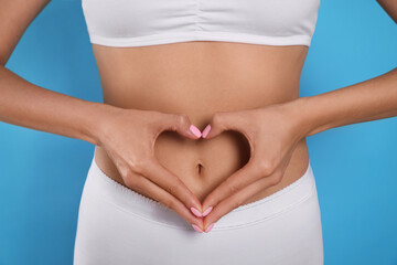 Woman in underwear making heart with hands on her belly against light blue background, closeup....