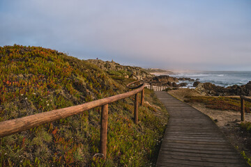 Fototapeta na wymiar Wooden walkway with hill with fence and carpobrotus edulis cactus, footpath in seascape at São Paio beach, cliffs and fog on horizon at evening, Labruge PORTUGAL