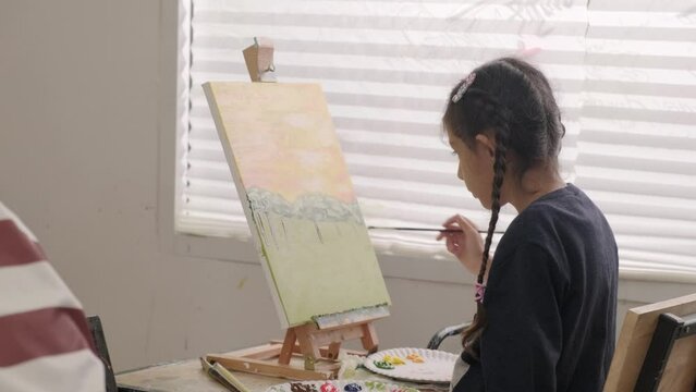 One little girl concentrates on acrylic color picture painting on canvas with multiracial kids in an art classroom, creative learning with talents and skills in the elementary school studio education.