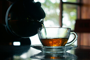 Pouring hot tea into a transparent cup by the window with a rustic blurred background.