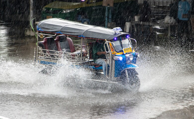 A traditional motorized tricycle - tuk tuk drives through a puddle on the road in heavy rain, Thailand - obrazy, fototapety, plakaty