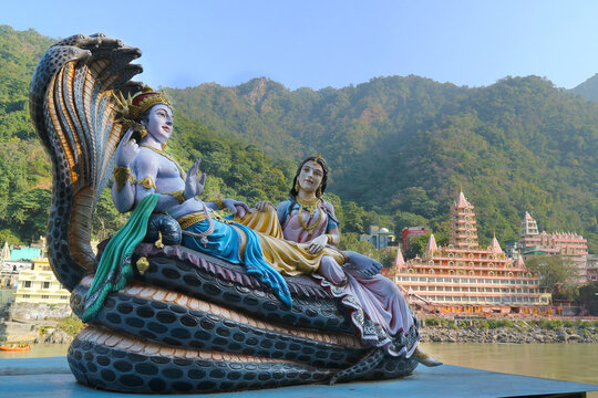 Sculpture of supreme god Vishnu reclining on the snake Ananta and Lakshmi massaging his feet on the banks of the Ganges in Rishikesh, India against the backdrop of temple and Lakshman Jhula bridge.
