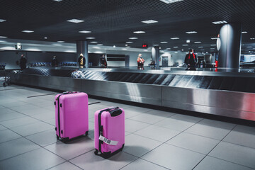 Selective focus on two pink luggage travel wheeled suitcases next to the baggage conveyor belt in an arrival zone of an airport terminal and people waiting for their bags in a defocused background