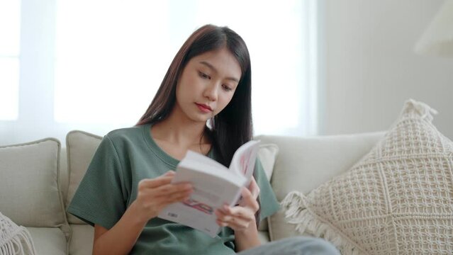 Happy young asian woman relax on comfortable couch at home, reading book and sitting on sofa