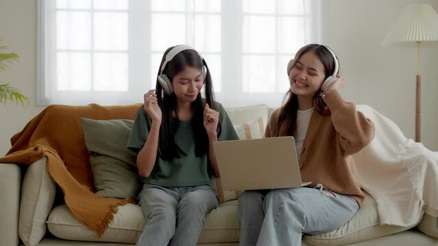 Two young asian women listening to music on couch in living room at home. Happy two asia female using computer laptop, wearing headset and sitting on sofa