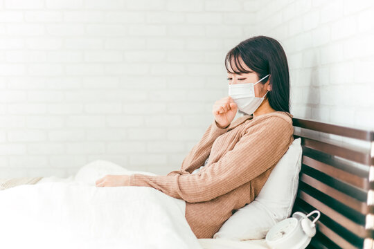 風邪をひいてベッドで咳をする体調不良のマスクをしたアジア人女性
