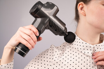 A caucasian woman uses a massager gun for pain in the muscles of her back and shoulders.