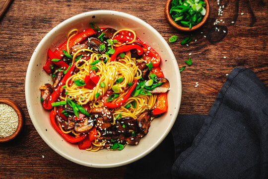 Vegan Stir fry noodles with vegetables, paprika, mushrooms, chives and sesame seeds in bowl. Wooden table background, top view, copy space