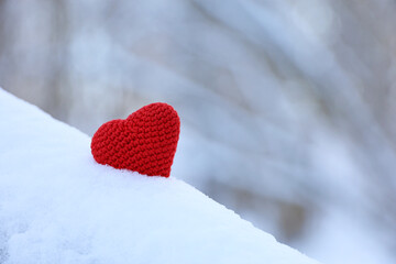 Fototapeta premium Red knitted heart in the snow on blurred forest background. Symbol of romantic love, Valentine's card