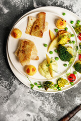 Top view baked sea fish fillet with vegetables. Tilapia fillet with broccoli, cherry tomatoes, fried potatoes, green peas and lettuce. Sea food on a white plate. Mediterranean Diet. Copy space
