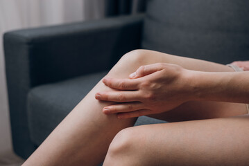 Closeup of unhealthy young woman sitting on sofa and feeling knee pain, doing massage knee at home. Female legs with meniscus injury or chronic joint inflammation. Healthcare and medical concept