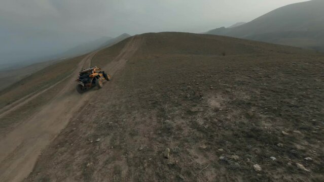 Yellow buggy car drives along brown ground road and jumps turning on meadow against hills silhouettes on horizon. Aerial cinematic fpv follow view on sport drone
