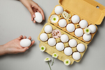 Female hands holds eggs on gray background with egg boxes with eggs and flowers