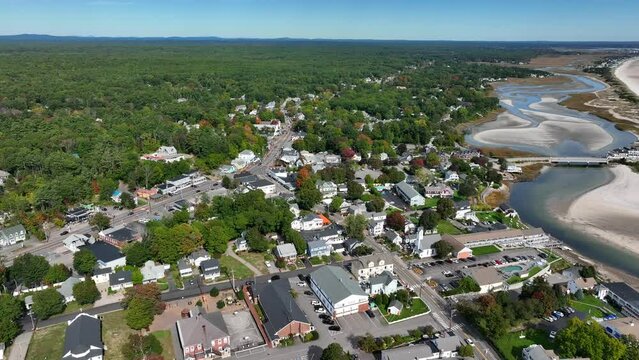 Aerial of New England beach town. Sandy shoreline with waves crossing and residential community. Forest surrounds village.