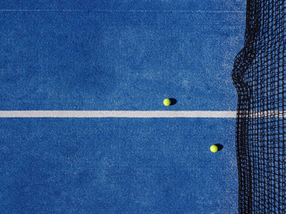 Overhead aerial view of two balls near the net of a blue paddle tennis court. Racket sports