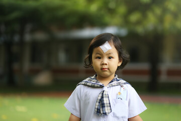 An Asian little girl who wearing a kindergarten uniform and has a wound sewn on her forehead 