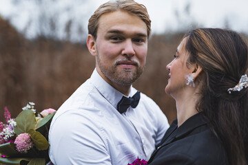 Proud groom - 30-year-old bearded blond Scandinavian man - looking at camera with a soft smile on his face while his beloved spouse is cuddling him. Blurred background. High quality photo