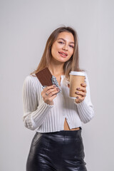 A beautiful young brown-haired woman eats a chocolate bar and drinks coffee on a white background. The girl is dressed in a white knitted blouse and leather pants