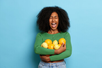 Indoor shot of emotional curly girl holding fresh oranges and lemons fruits, keeps mouth widely opened, dressed in casual cloth