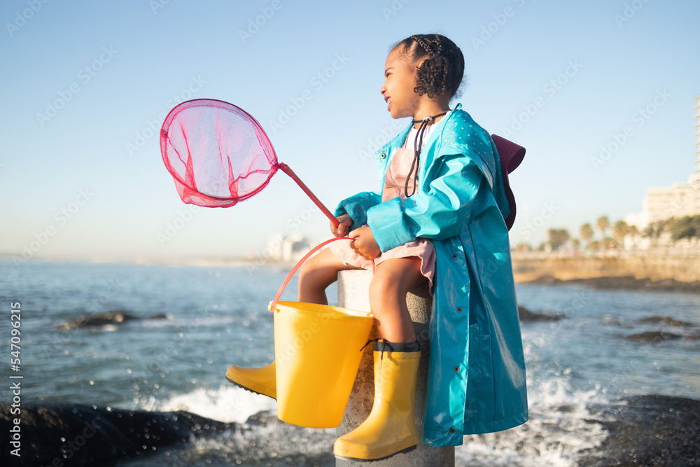 Wall mural Girl, beach and fishing net with bucket, water and rock pool for outdoor adventure with boots, smile and ocean. Black child, sea and happy for fish, waves or sea in nature, learning and sunshine