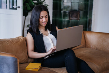 Young purposeful African American businesswoman in black suit and white blouse sits on cozy couch with laptop, typing, makes video call toothy smiles. Pretty Brazilian student girl at distant lesson.