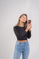 A beautiful brown-haired girl holds a chocolate bar on a white background. Young brunette woman wearing a black blouse and blue jeans