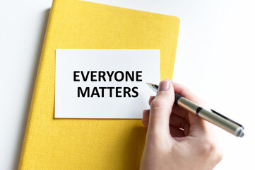 EVERYONE MATTERS text on a card on a yellow notebook on a light background next to a person's hand with a pen